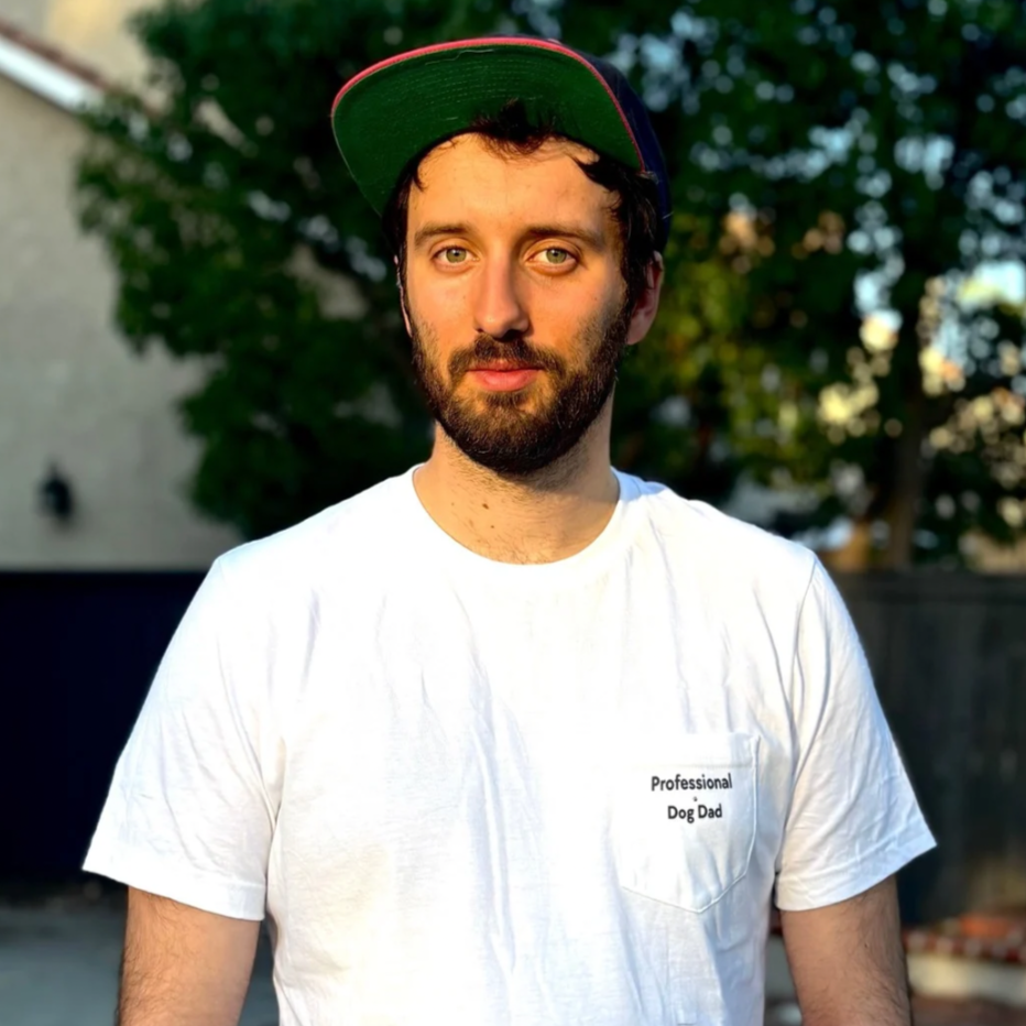 A man with a hat on and a white tee shirt that says professional dog dad on the front pocket. He's outside and looking at the camera. Kimberly's Canine Care t-shirt for dog fathers day.