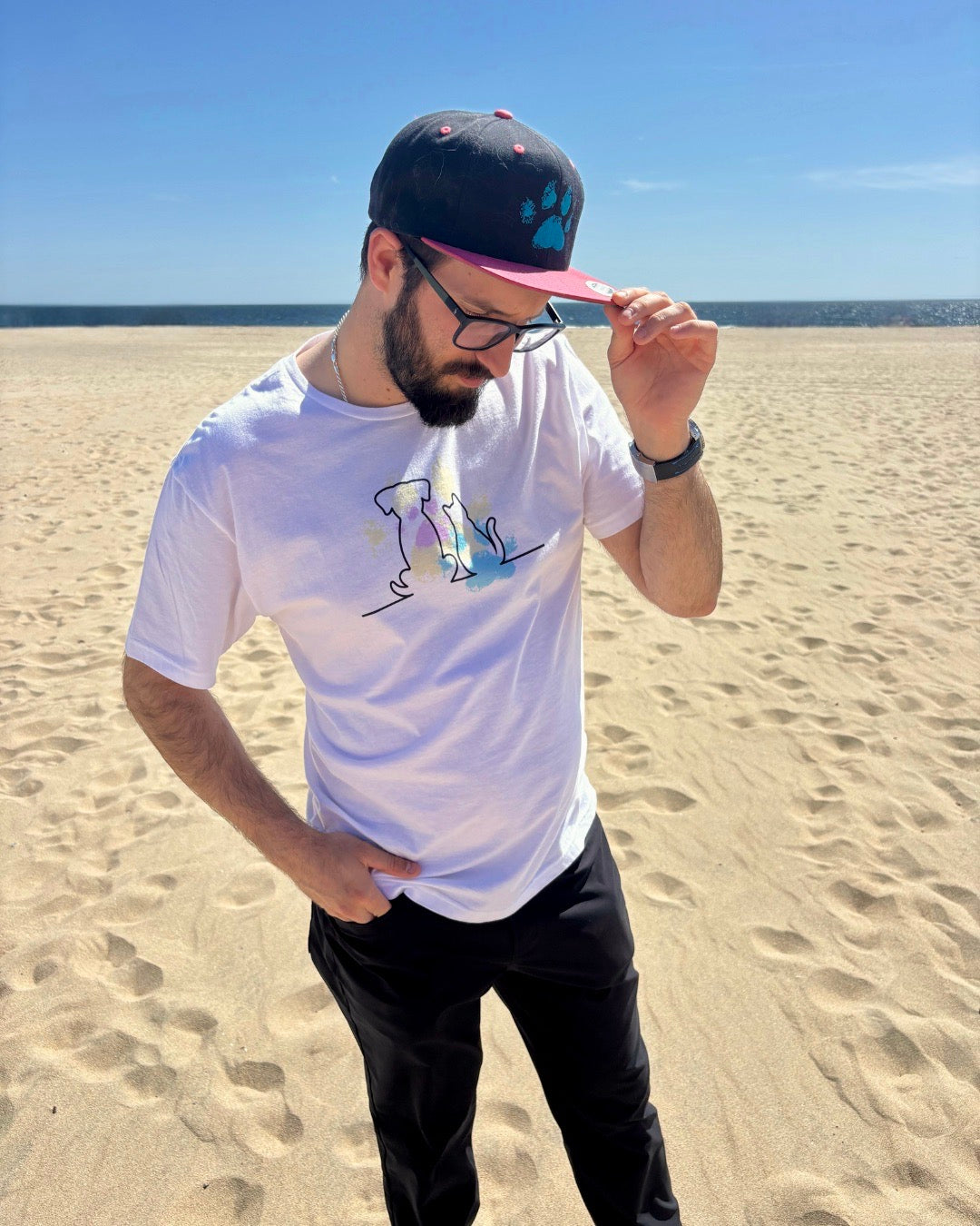 man on the beach, with the ocean in the background. Showcasing his black and pink hat with a blue dog paw print on it. 