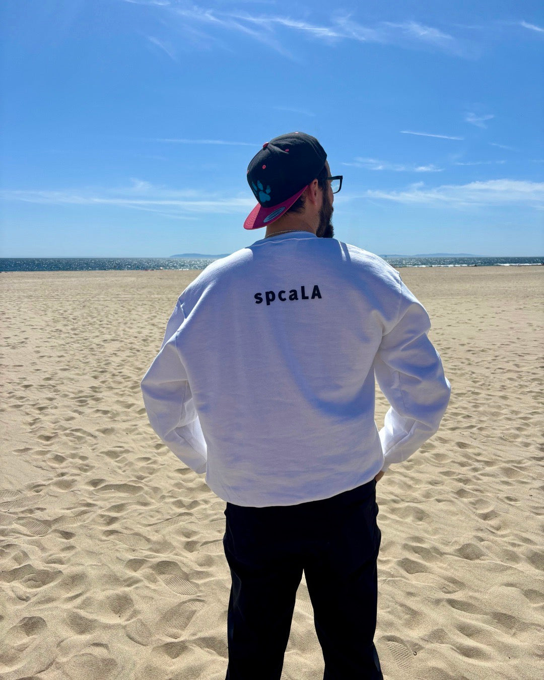 man on beach with hat. Ocean in the background. He's showcasing his white sweatshirt that says spcaLA on the back.