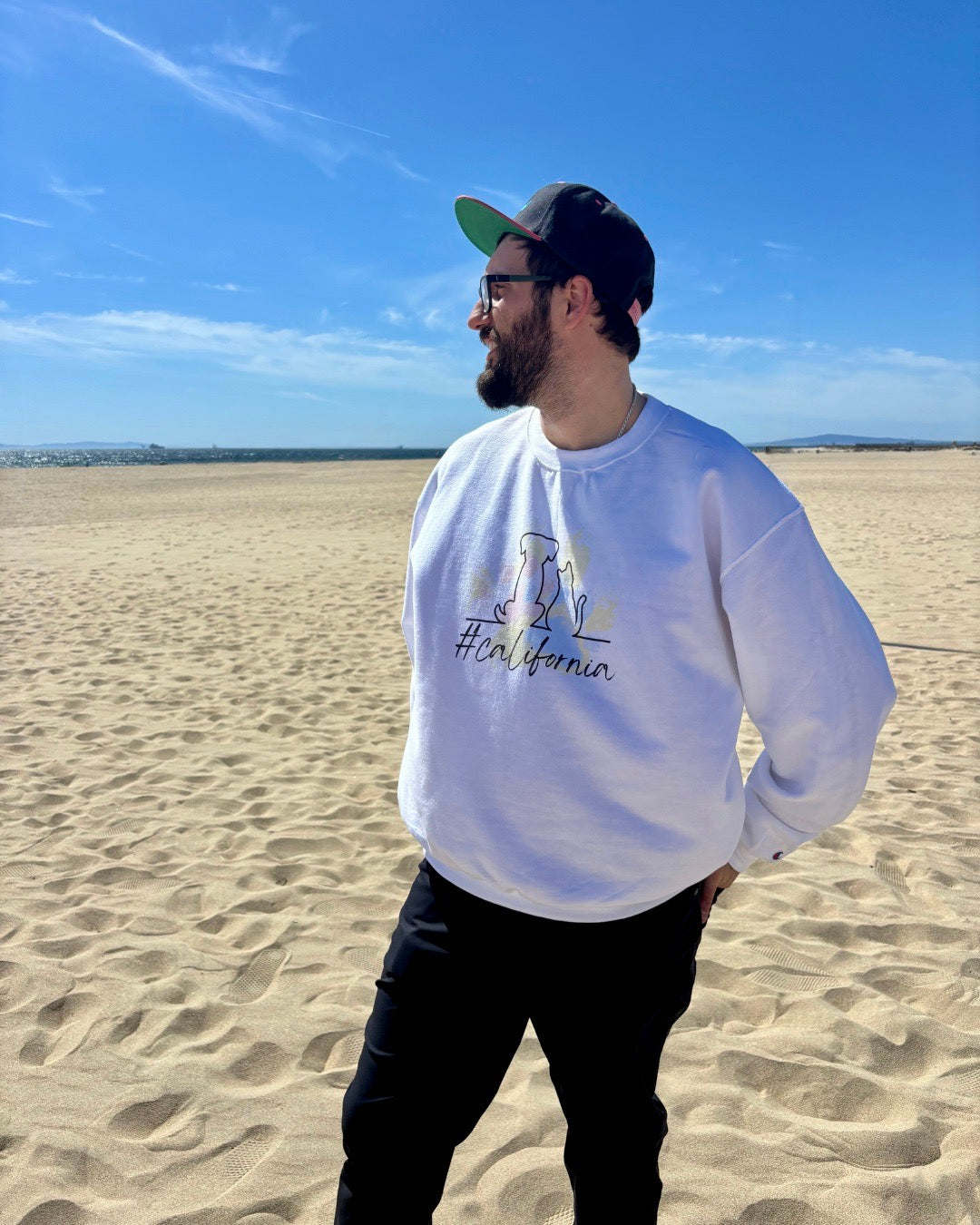 Side view of a man on beach with hat smiling. Ocean in the background. He's showcasing his white sweatshirt that says #california and has  cat and dog silhouette with colorful pawprints in the background. 