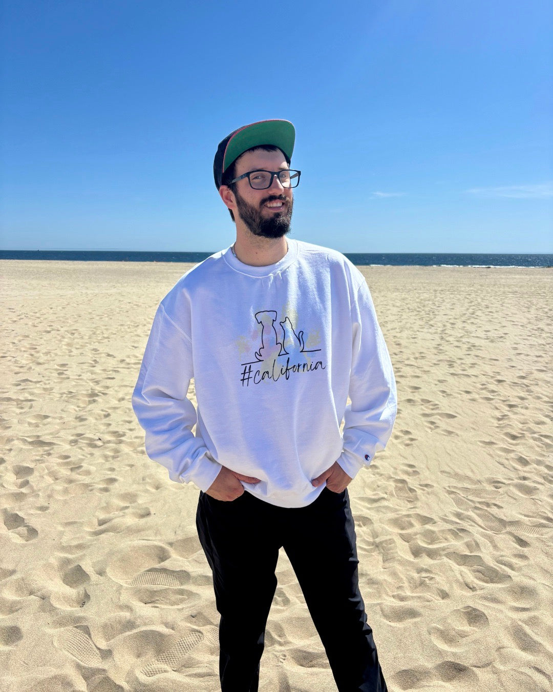 man on beach with hat smiling. Ocean in the background. He's showcasing his white sweatshirt that says #california and has cat and dog silhouette with colorful pawprints in the background. 