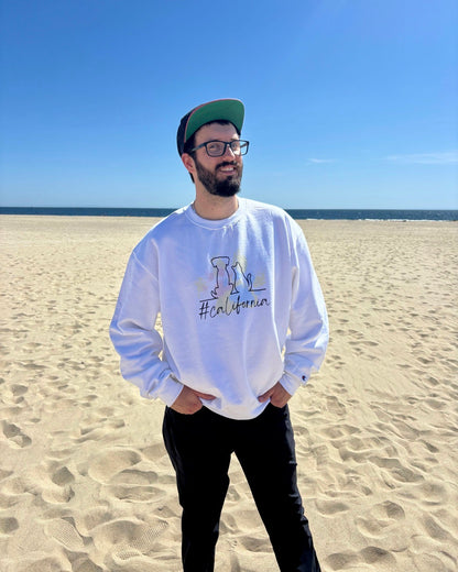 man on beach with hat smiling. Ocean in the background. He's showcasing his white sweatshirt that says #california and has cat and dog silhouette with colorful pawprints in the background. 
