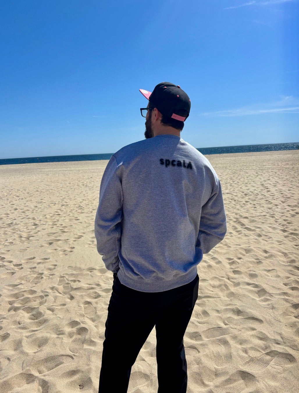 man on beach with hat. Facing the ocean in the background. He's showcasing his sweatshirt that says spcaLA on the back. 