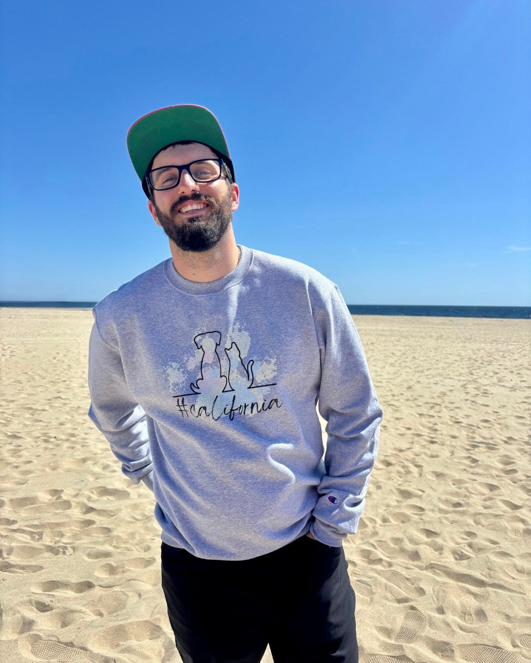 man on beach with hat laughing. Ocean in the background. He's showcasing his sweatshirt that says #california and has  cat and dog silhouette with colorful pawprints in the background. 